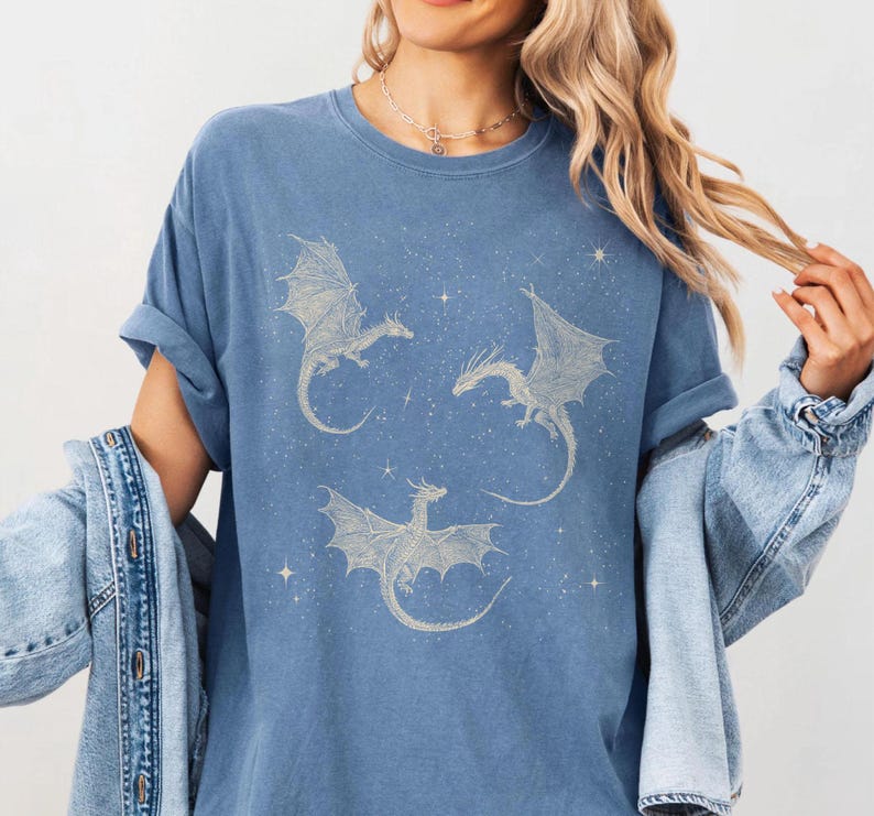 K&ouml;nnte beinhalten: Blaues T-Shirt mit einem Himmelsdesign, das drei detaillierte Drachenillustrationen zeigt. Die Drachen haben ausgebreitete Fl&uuml;gel und sind von sternartigen Akzenten umgeben. Das Shirt hat eine lockere Passform und gerollte &Auml;rmel.
