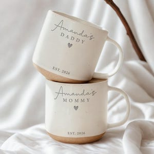 May include: Two white ceramic mugs with a speckled finish, stacked on top of each other. Each mug has a tan base and a curved handle. The top mug reads "Amanda's DADDY" with a heart and "EST. 2026". The bottom mug reads "Amanda's MOMMY" with a heart and "EST. 2026".