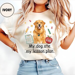 Puede incluir: Una camiseta de color marfil con un gr&aacute;fico de un perro golden retriever rodeado de material escolar, como un l&aacute;piz, un cray&oacute;n verde, un portapapeles y una manzana roja con la etiqueta Ozzy. El texto dice My dog ate my lesson plan.
