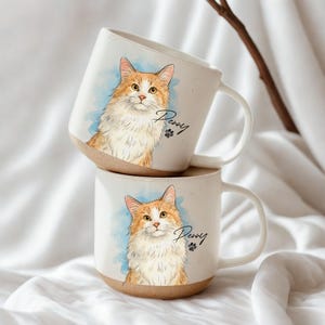 May include: Two white ceramic mugs stacked, each with a watercolour illustration of a fluffy ginger and white cat. The cat's name, "Penny", is written in script below the image, along with a paw print. The base of the mugs are a natural tan colour.