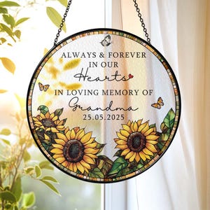 May include: A round, glass memorial suncatcher with a black metal frame and chain. The suncatcher features sunflowers, butterflies, and the text "ALWAYS & FOREVER IN OUR HEARTS IN LOVING MEMORY OF Grandma 25.05.2025".
