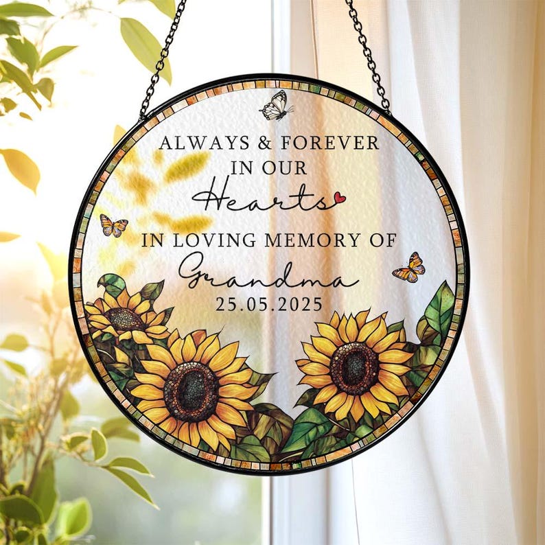 May include: A round, glass memorial suncatcher with a black metal frame and chain. The suncatcher features sunflowers, butterflies, and the text "ALWAYS & FOREVER IN OUR HEARTS IN LOVING MEMORY OF Grandma 25.05.2025".