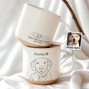 May include: Two white ceramic mugs with brown bottoms. One mug has the text "BEST DOG DAD EVER EST. 2026" and a paw-shake graphic. The other mug has a dog portrait and the name "Cookie". A photo of a dog is included.