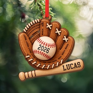 May include: A wooden baseball-themed ornament featuring a brown glove with white lacing, a light-colored baseball with the text 2026, and a horizontal baseball bat engraved with the name LUCAS. A red ribbon is attached to the top for hanging.