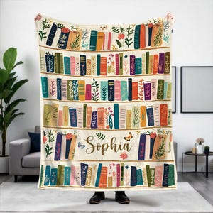 May include: A white fleece blanket with a bookshelf design featuring colorful books, green plants, and butterflies. The name "Sophia" is printed in a decorative font on the blanket.