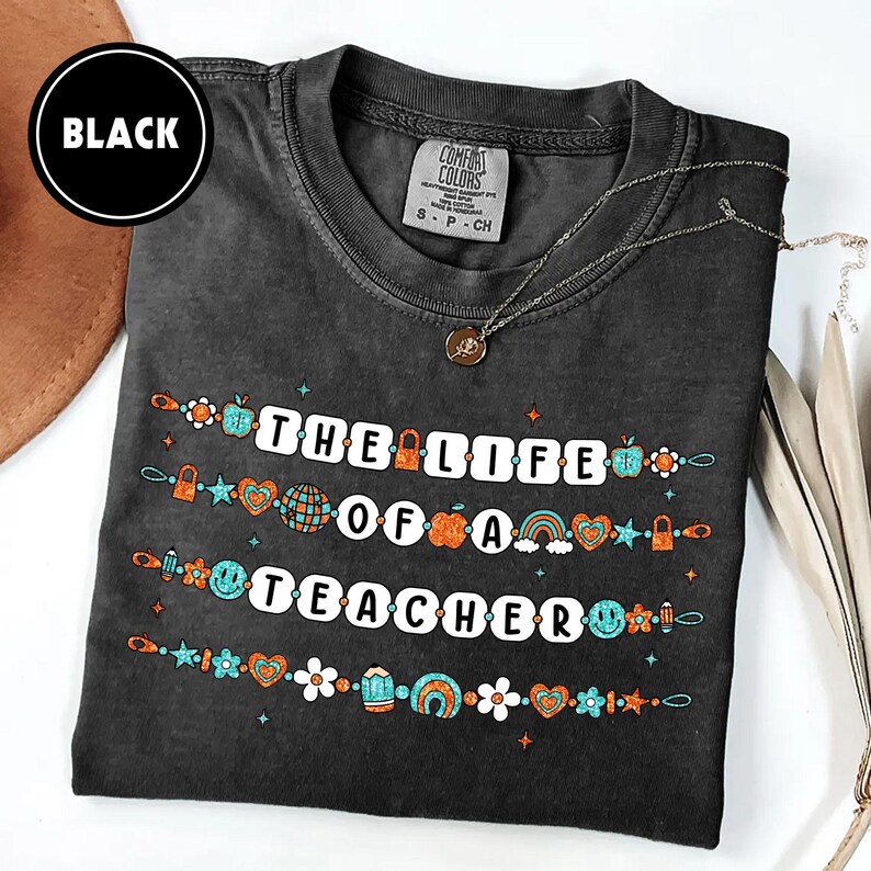 May include: Black t-shirt with the text "THE LIFE OF A TEACHER" in white block letters. The text is decorated with colorful illustrations of apples, hearts, and rainbows. A silver necklace with a pendant is also visible.