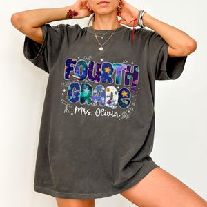 May include: Dark gray t-shirt with a colorful "Fourth Grade Mrs. Olivia" graphic. The design features a space theme with stars, planets, and a rocket. The text is in a playful, multi-colored font.