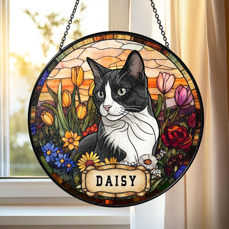 May include: A round stained glass window featuring a black and white cat named Daisy, surrounded by colorful flowers. The cat is the focal point, set against a backdrop of a sunset. The window is framed with a black metal border and hangs from a chain.