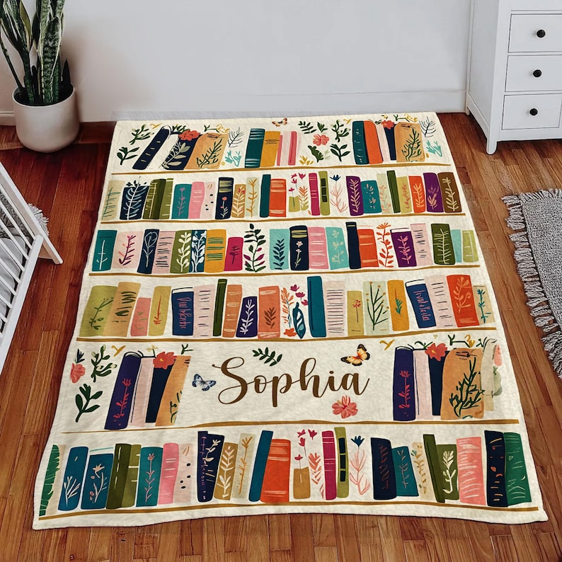 May include: A cream-colored throw blanket with a bookshelf design featuring colorful books, green leaves, and butterflies. The name "Sophia" is written in gold lettering on the blanket.