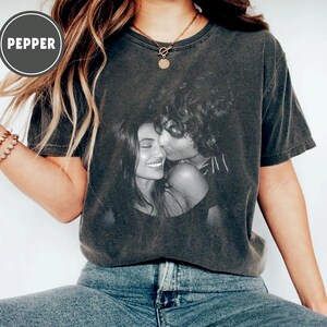 May include: Dark grey t-shirt with a black and white photograph of a couple kissing. The shirt has a faded, vintage appearance. The word "PEPPER" is printed in a circle on the upper left side. The model is wearing blue jeans.