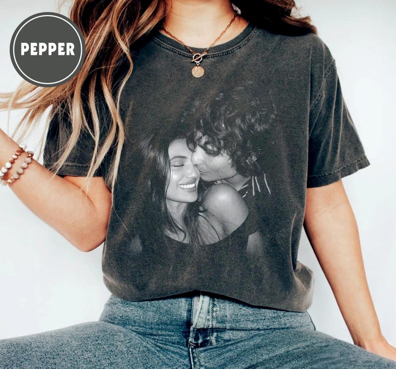 May include: Dark grey t-shirt with a black and white photograph of a couple kissing. The shirt has a faded, vintage appearance. The word "PEPPER" is printed in a circle on the upper left side. The model is wearing blue jeans.