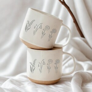 May include: Two cream-coloured ceramic mugs stacked on top of each other, each with a light brown base. The mugs are decorated with a grey floral design. These mugs have a simple, elegant design, ideal for tea or coffee.
