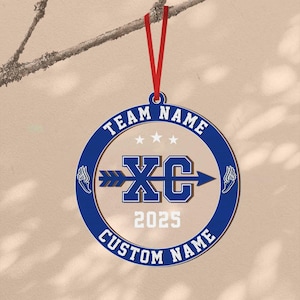 Puede incluir: Adorno del equipo XC azul y blanco con una cinta roja para colgar. El adorno presenta el texto "TEAM NAME" y "CUSTOM NAME" alrededor del borde, con "XC" y "2025" en el centro. Gr&aacute;ficos de pies alados a ambos lados.
