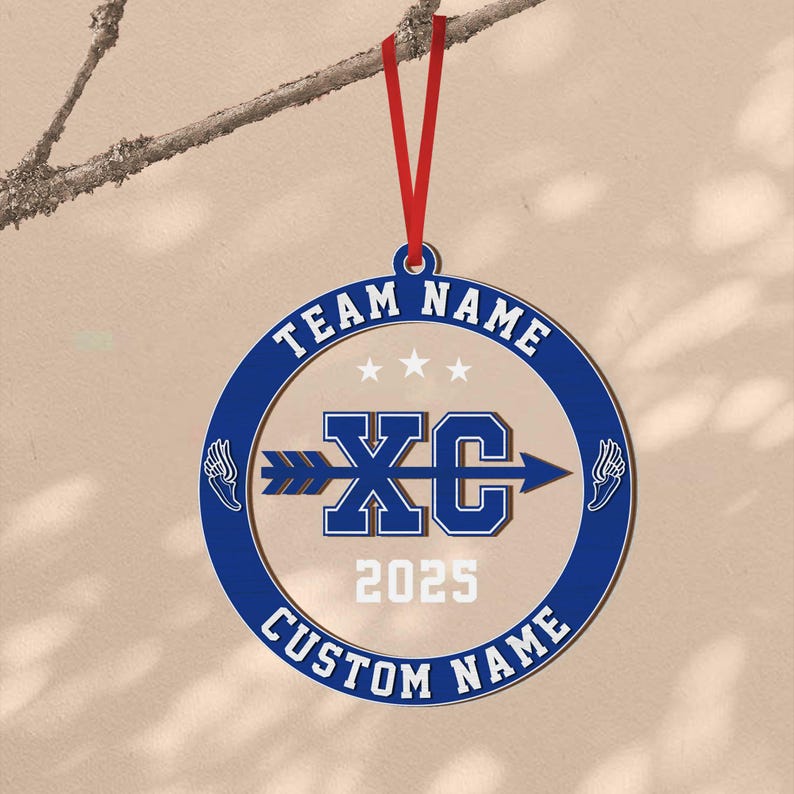 Puede incluir: Adorno del equipo XC azul y blanco con una cinta roja para colgar. El adorno presenta el texto "TEAM NAME" y "CUSTOM NAME" alrededor del borde, con "XC" y "2025" en el centro. Gr&aacute;ficos de pies alados a ambos lados.