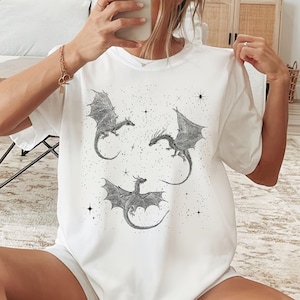 K&ouml;nnte beinhalten: Wei&szlig;es T-Shirt mit einer Schwarz-Wei&szlig;-Illustration von drei Drachen mit ausgebreiteten Fl&uuml;geln und Sternen. Die Drachen sind detailliert mit Schuppen und Krallen. Ein l&auml;ssiges Kleidungsst&uuml;ck f&uuml;r den Alltag.