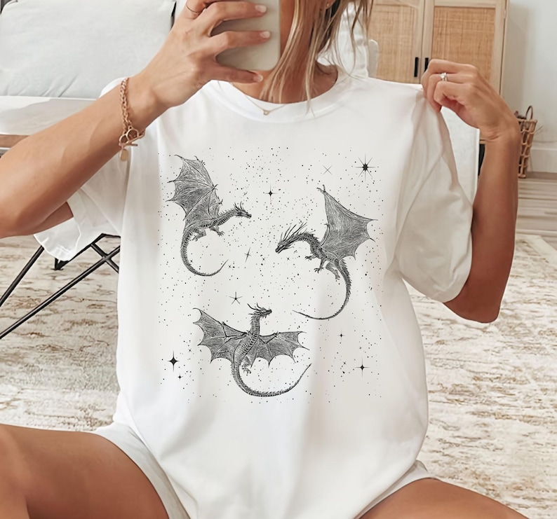 K&ouml;nnte beinhalten: Wei&szlig;es T-Shirt mit einer Schwarz-Wei&szlig;-Illustration von drei Drachen mit ausgebreiteten Fl&uuml;geln und Sternen. Die Drachen sind detailliert mit Schuppen und Krallen. Ein l&auml;ssiges Kleidungsst&uuml;ck f&uuml;r den Alltag.