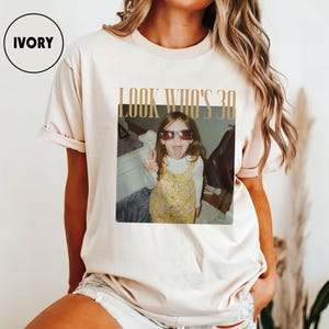 Może przedstawiać: Kremowa koszulka z nadrukiem "LOOK WHO'S 30". Grafika przedstawia zdjęcie małej dziewczynki w okularach przeciwsłonecznych, pokazującej język. Dziewczynka ma na sobie ż&oacute;łtą sukienkę w kwiaty.