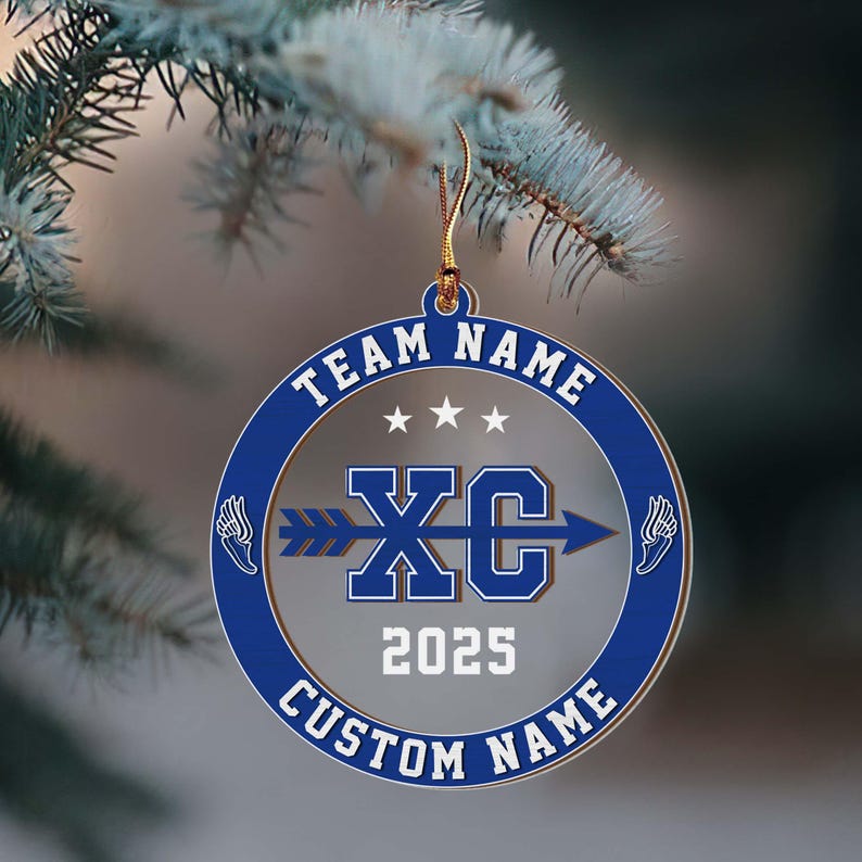 Puede incluir: Un adorno redondo azul y plateado con los textos "TEAM NAME" y "CUSTOM NAME". El centro presenta "XC" con una flecha, el a&ntilde;o "2025" y dos dise&ntilde;os de alas. Una cuerda dorada est&aacute; unida para colgarlo.