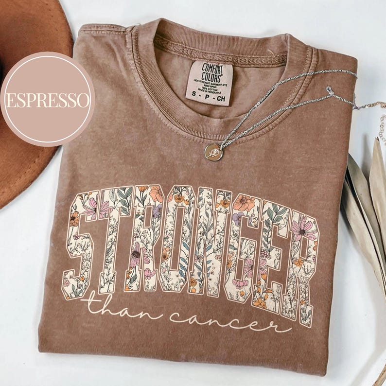 May include: Espresso-colored t-shirt with the words "STRONGER than cancer" in a floral design. The shirt has a crew neck and short sleeves. A silver necklace with a pendant is draped across the shirt.