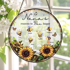 May include: A round, decorative glass sign with a black metal chain for hanging. The sign features the text "Nana's reasons to Bee happy" with cartoon bees and names. Sunflowers and butterflies decorate the bottom and sides.