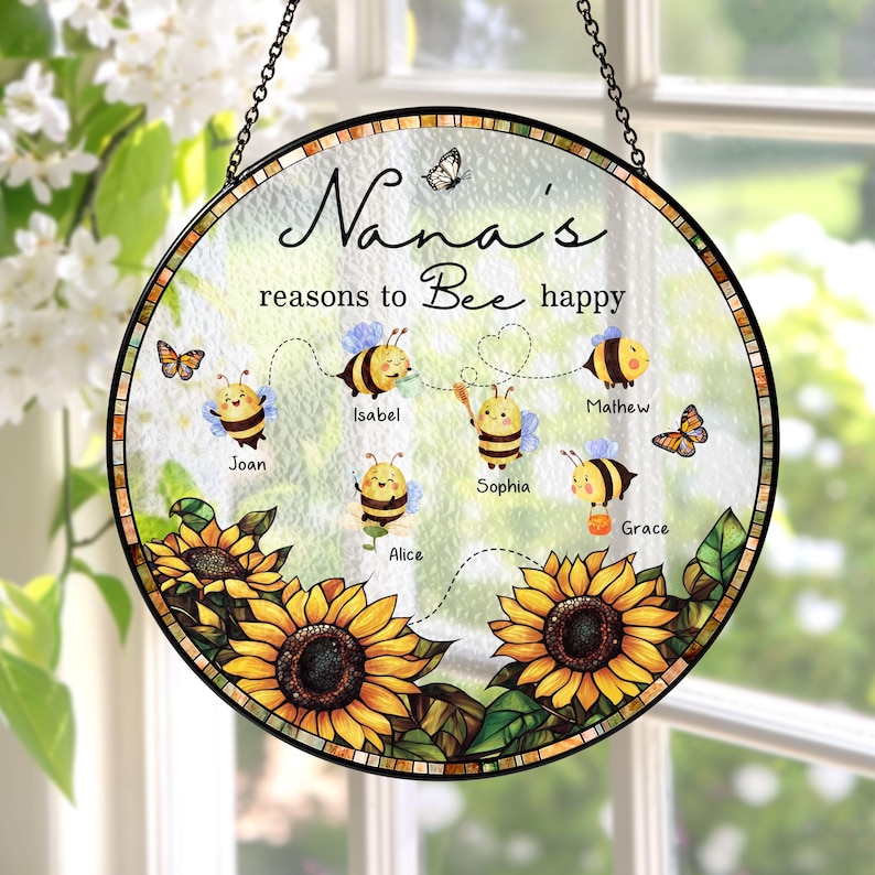 May include: A round, decorative glass sign with a black metal chain for hanging. The sign features the text "Nana's reasons to Bee happy" with cartoon bees and names. Sunflowers and butterflies decorate the bottom and sides.