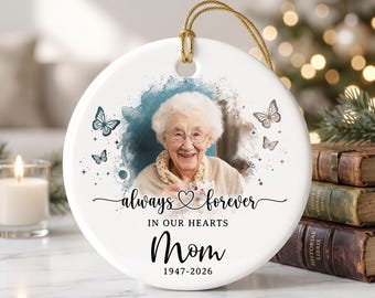 Op de afbeelding: Een rond wit keramieken herdenkingsornament met een portret van een oudere vrouw met een bril, omringd door blauwe aquarelaccenten en vlinders. De tekst luidt "always forever IN OUR HEARTS Mom 1947-2026" met een gouden ophangkoord.