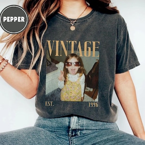 Puede incluir: Camiseta gris oscuro con un dise&ntilde;o vintage. La camiseta presenta la palabra "VINTAGE" en fuente dorada, junto con una foto de una ni&ntilde;a con gafas de sol y sacando la lengua. El texto "EST. 1996" est&aacute; debajo de la foto.
