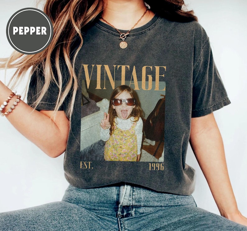 Puede incluir: Camiseta gris oscuro con un dise&ntilde;o vintage. La camiseta presenta la palabra "VINTAGE" en fuente dorada, junto con una foto de una ni&ntilde;a con gafas de sol y sacando la lengua. El texto "EST. 1996" est&aacute; debajo de la foto.