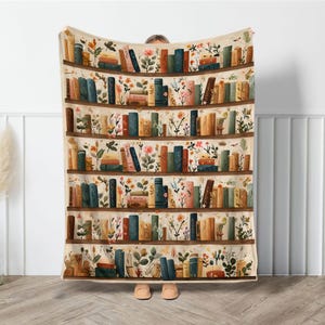May include: A beige throw blanket featuring a whimsical bookshelf design. The blanket showcases rows of books in various colors, interspersed with floral and botanical illustrations. The overall design is a cozy, artistic representation of a library.
