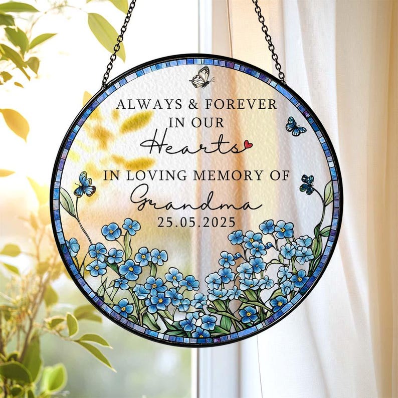 May include: Round stained glass memorial with the text "ALWAYS & FOREVER in our Hearts, IN LOVING MEMORY OF Grandma 25.05.2025". Features blue flowers, butterflies, and a black chain for hanging. The glass has a blue and white border.