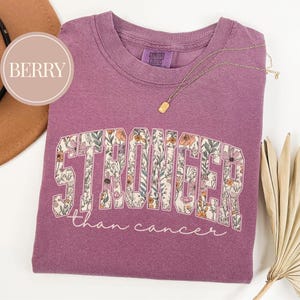 May include: Berry-colored t-shirt with the word "STRONGER" in a floral design, and the phrase "than cancer" in script. A gold necklace with a rectangular pendant rests on the shirt. The shirt has a Comfort Colors tag.
