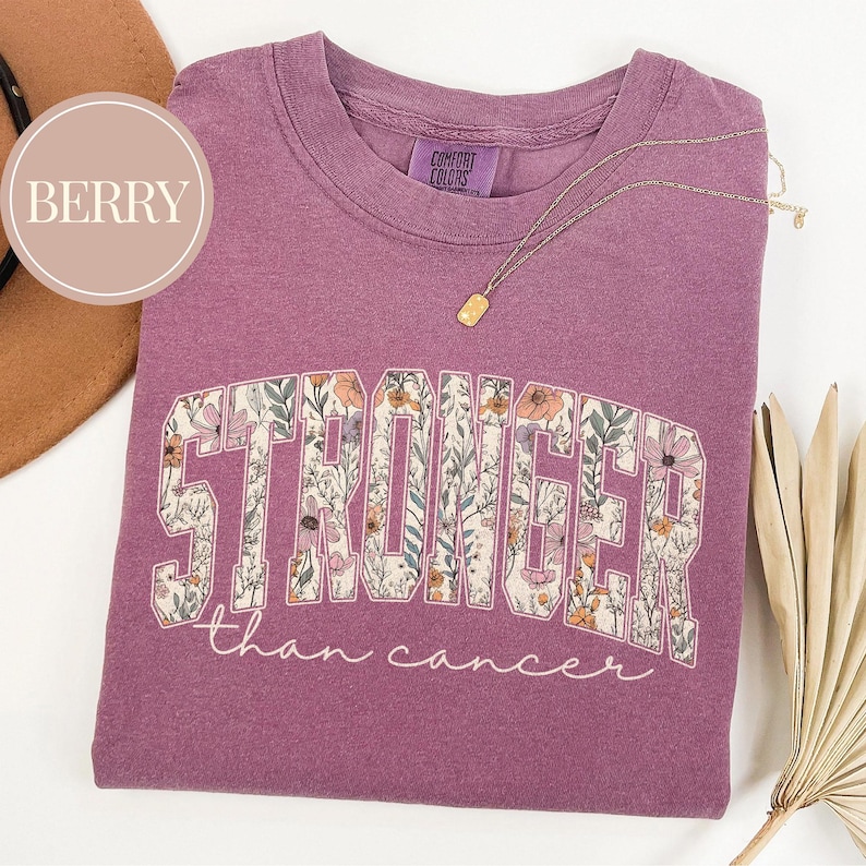 May include: Berry-colored t-shirt with the word "STRONGER" in a floral design, and the phrase "than cancer" in script. A gold necklace with a rectangular pendant rests on the shirt. The shirt has a Comfort Colors tag.