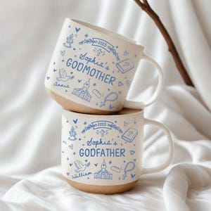 May include: Two white ceramic mugs with blue illustrations and text. One mug says "Sophia's GODMOTHER" and the other says "Sophia's GODFATHER". Both mugs have a light brown base and a white handle. The mugs are decorated with religious symbols.