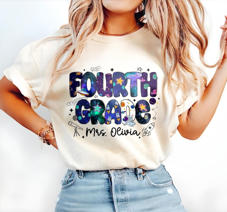 May include: Cream-colored t-shirt featuring the words "FOURTH GRADE" in a colorful, space-themed design. The design includes planets, stars, and a rocket ship. Below the text is "Mrs. Olivia".