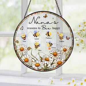 May include: Round glass hanging decoration with a black chain. The design features a floral border with daisies and the text "Nana's reasons to Bee happy." Includes cartoon bees with names like Joan and Isabel.