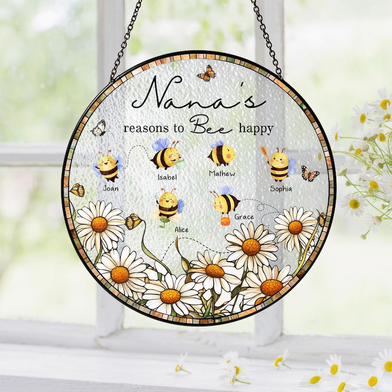 May include: Round glass hanging decoration with a black chain. The design features a floral border with daisies and the text "Nana's reasons to Bee happy." Includes cartoon bees with names like Joan and Isabel.