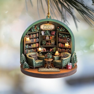 May include: A decorative hanging ornament featuring a miniature bookstore interior with two green patterned armchairs, a small round table, and shelves filled with books. A sign above the shelves reads Amelia's Bookstore, with small lamps and trees.