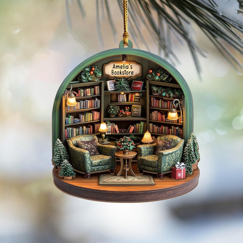 May include: A decorative hanging ornament featuring a miniature bookstore interior with two green patterned armchairs, a small round table, and shelves filled with books. A sign above the shelves reads Amelia's Bookstore, with small lamps and trees.