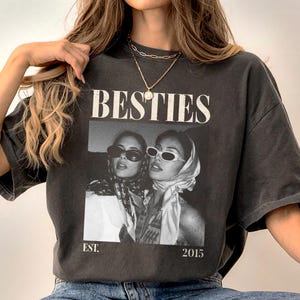 May include: Dark gray t-shirt featuring a black and white image of two women wearing sunglasses and headscarves. The word "BESTIES" is printed above the image, with "EST. 2015" below. The shirt has short sleeves.