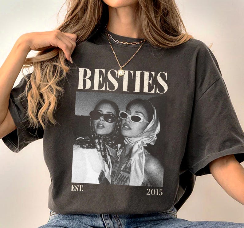May include: Dark gray t-shirt featuring a black and white image of two women wearing sunglasses and headscarves. The word "BESTIES" is printed above the image, with "EST. 2015" below. The shirt has short sleeves.