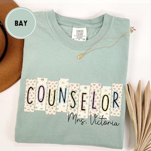 May include: A light green t-shirt with the word "COUNSELOR" in a floral design, with "Mrs. Victoria" below. The shirt has a crew neck and short sleeves. A gold necklace and a brown hat are also visible.