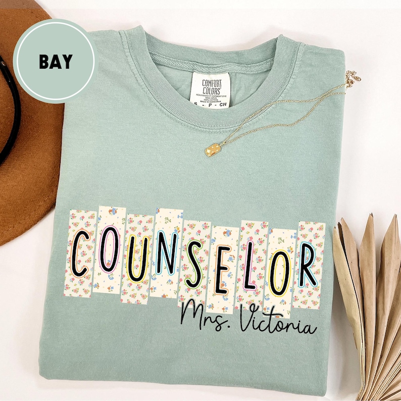 May include: A light green t-shirt with the word "COUNSELOR" in a floral design, with "Mrs. Victoria" below. The shirt has a crew neck and short sleeves. A gold necklace and a brown hat are also visible.