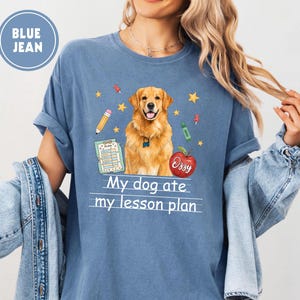 Puede incluir: Una camiseta color azul vaquero con un gr&aacute;fico de un perro golden retriever, un l&aacute;piz, un portapapeles, una manzana roja con el nombre Ozzy y estrellas amarillas. El texto My dog ate my lesson plan est&aacute; impreso en blanco bajo el perro.