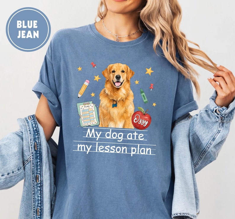 Puede incluir: Una camiseta color azul vaquero con un gr&aacute;fico de un perro golden retriever, un l&aacute;piz, un portapapeles, una manzana roja con el nombre Ozzy y estrellas amarillas. El texto My dog ate my lesson plan est&aacute; impreso en blanco bajo el perro.