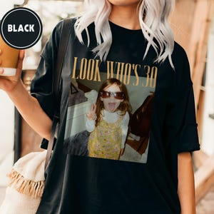 Może przedstawiać: Czarny t-shirt z nadrukiem zdjęcia. Zdjęcie przedstawia małą dziewczynkę w okularach przeciwsłonecznych z wystawionym językiem, z napisem "LOOK WHO'S 30" złotymi literami. Koszulka ma luźny kr&oacute;j.