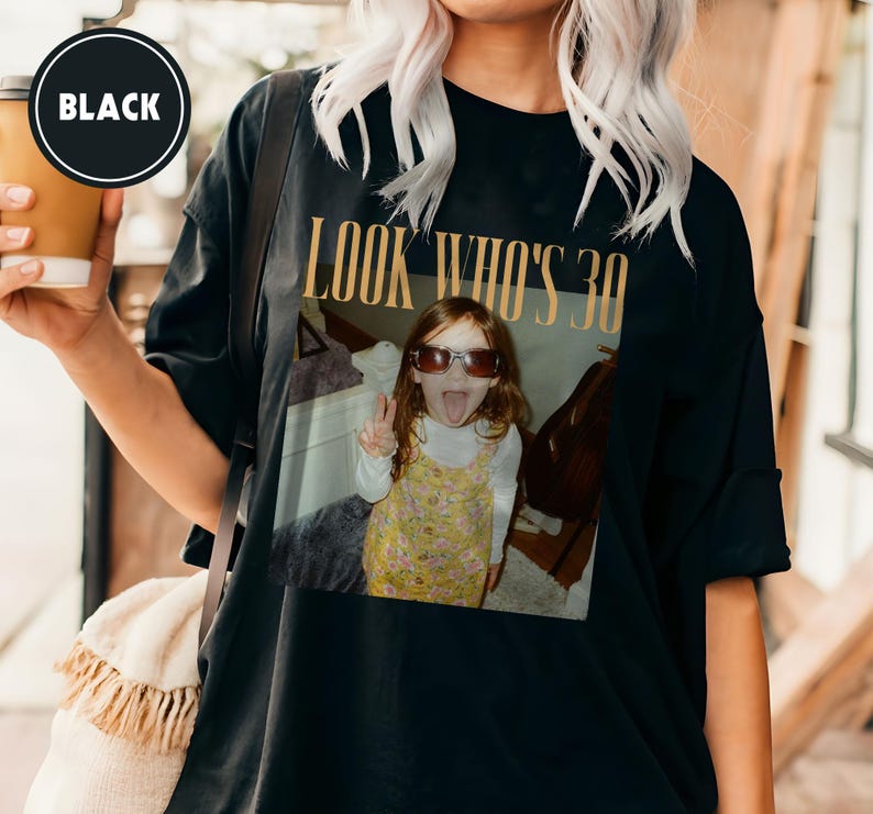 Może przedstawiać: Czarny t-shirt z nadrukiem zdjęcia. Zdjęcie przedstawia małą dziewczynkę w okularach przeciwsłonecznych z wystawionym językiem, z napisem "LOOK WHO'S 30" złotymi literami. Koszulka ma luźny kr&oacute;j.