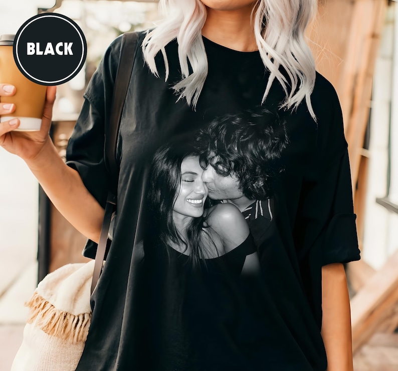 May include: Black oversized t-shirt with a black and white image of a couple kissing. The t-shirt has a round neckline and short sleeves. A black circle with the word "BLACK" is in the upper left corner.