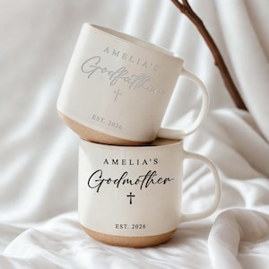 May include: Two stacked white ceramic mugs with a tan base. The top mug says "AMELIA'S Godfather" with a cross and "EST. 2026". The bottom mug says "AMELIA'S Godmother" with a cross and "EST. 2026".