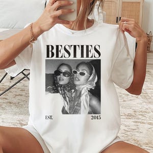 May include: White t-shirt with the word "BESTIES" in a bold, black font. Below the text is a black and white photo of two women wearing sunglasses and scarves. The text "EST. 2015" is printed below the photo.