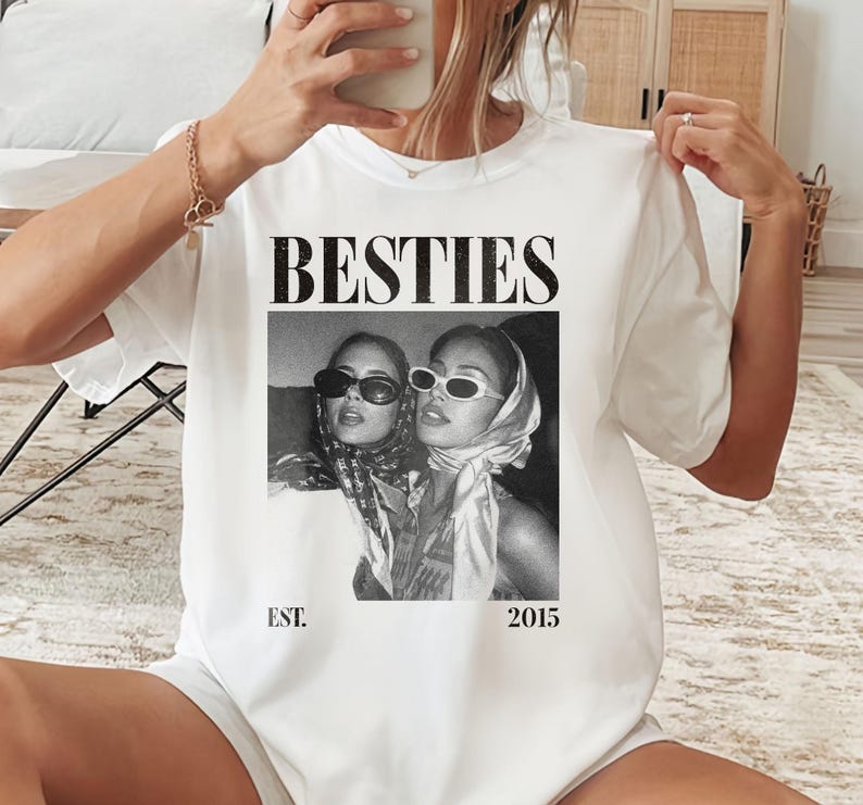 May include: White t-shirt with the word "BESTIES" in a bold, black font. Below the text is a black and white photo of two women wearing sunglasses and scarves. The text "EST. 2015" is printed below the photo.